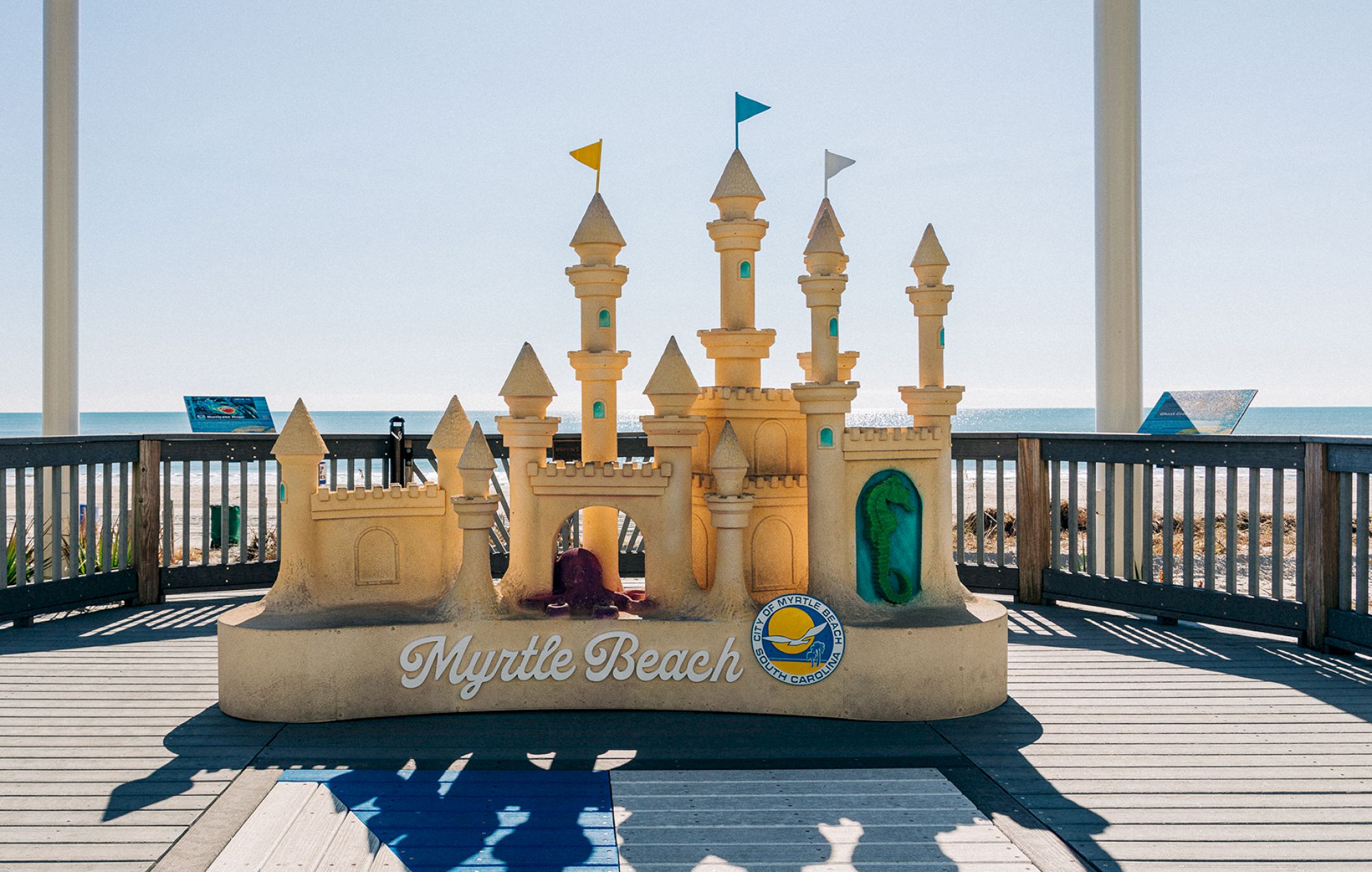 A sandcastle sculpture with "Myrtle Beach" written at the front, placed on a wooden deck with the ocean in the background.