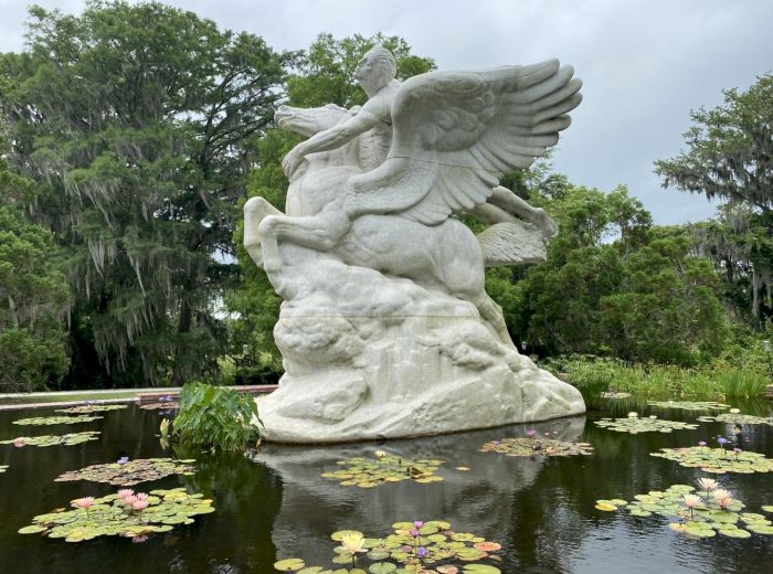 A large statue of a winged horse with a rider stands in a pond with water lilies surrounded by lush, green trees and foliage, creating a serene scene.