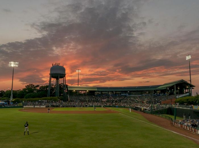 A baseball game is taking place at dusk, with a water tower and stadium lights in the background, under a dramatic sunset sky.