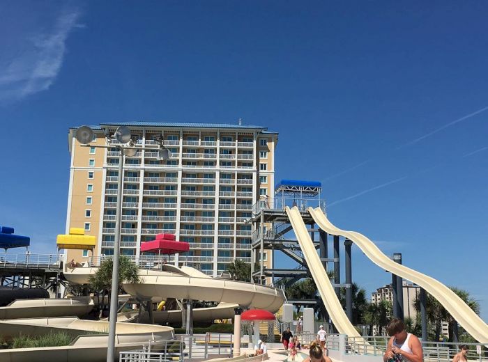 The image shows a water park with slides, a pool, and people in swimwear. A tall building is in the background under a clear blue sky.
