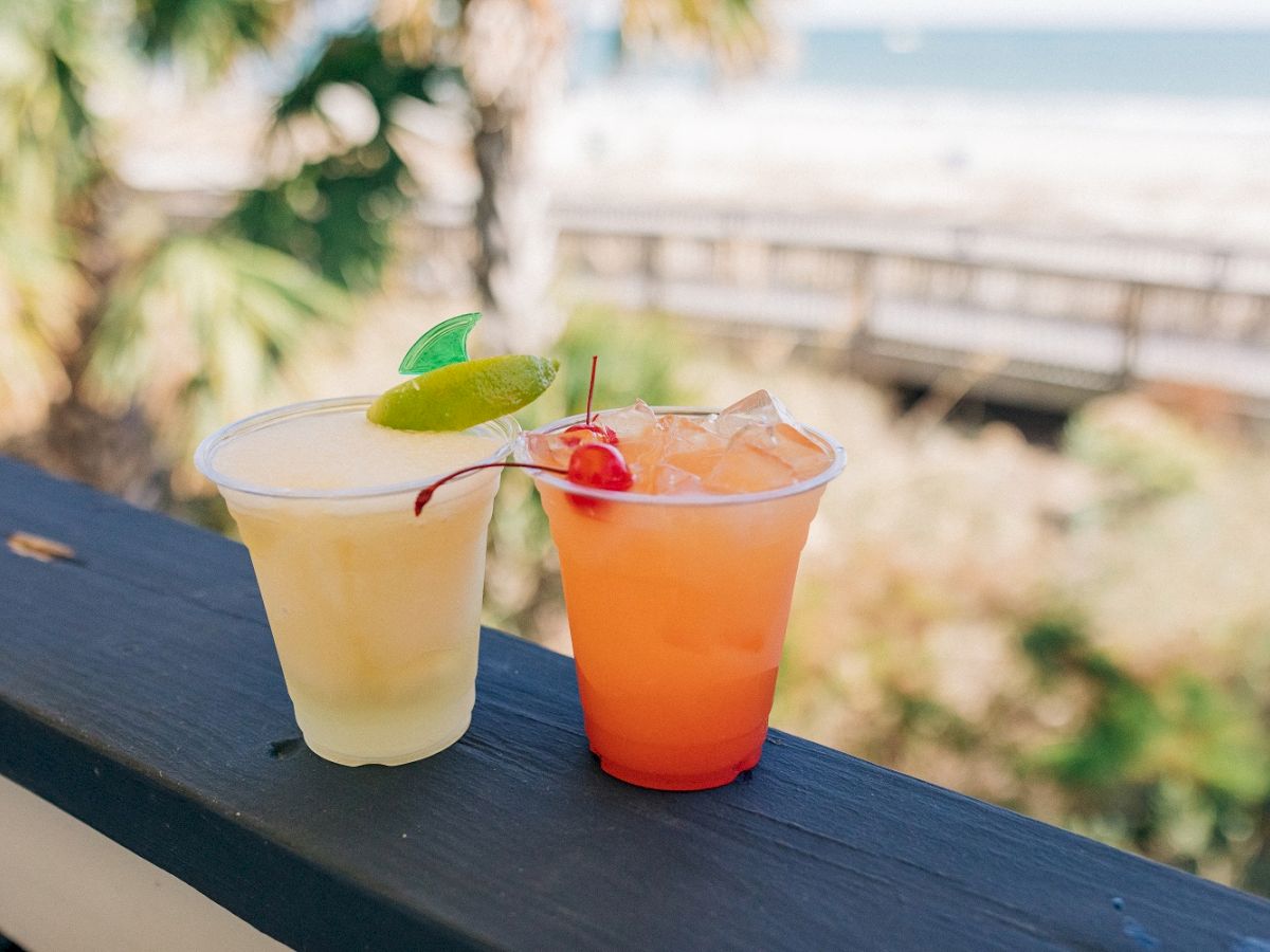 Two tropical drinks, one yellow with a lime garnish and one orange with a cherry garnish, placed on a wooden ledge with a beach view in the background.