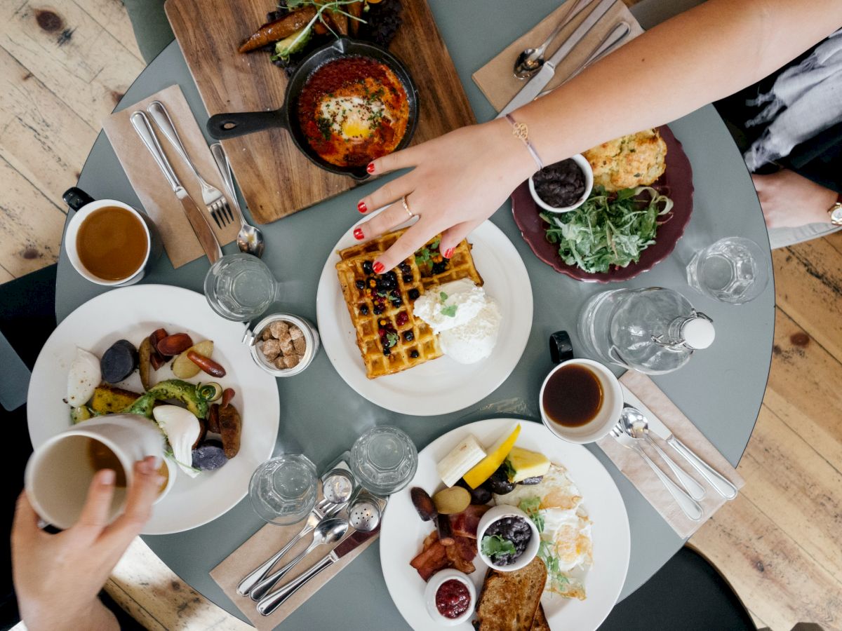 A top view of a dining table with waffles, fruits, vegetables, eggs, utensils, drinks, and hands reaching for food from different plates.