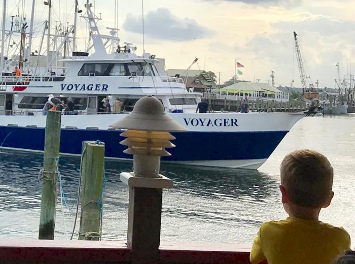 A child gazes at a large boat named "Voyager" docked at a marina, with a backdrop of cloudy skies and other vessels.