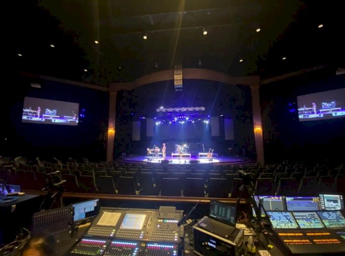 A concert hall is shown from the sound and lighting control booth, with a stage set up and large screens on either side, featuring live performers.