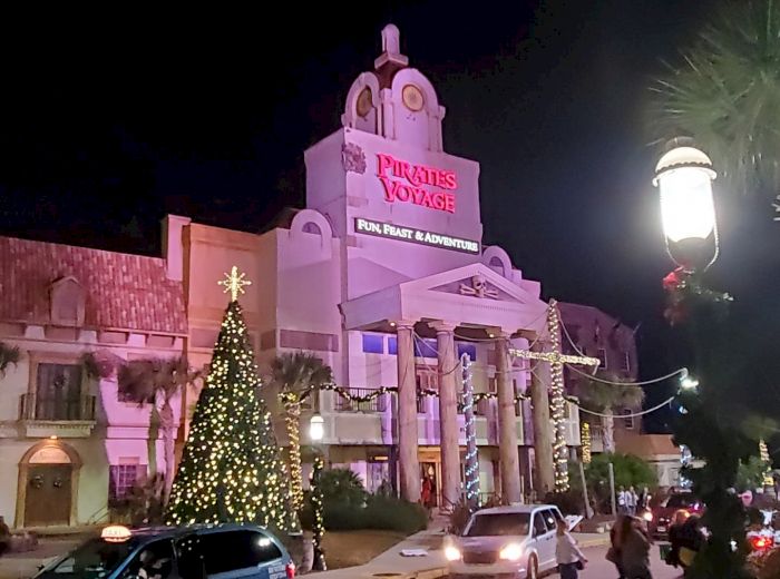This image shows the exterior of "Pirates Voyage" with festive decorations, including a lit Christmas tree, cars, and people walking at night.