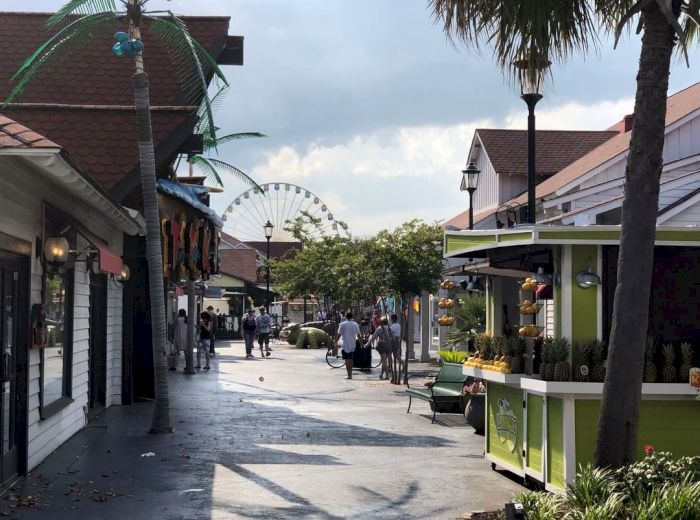A pedestrian-friendly outdoor area with shops, palm trees, people, and a Ferris wheel in the background.