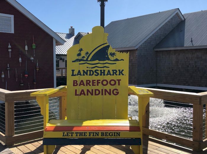 A large yellow chair with "Landshark Barefoot Landing" and "Let the Fin Begin" text, located on a wooden deck near buildings and a waterfront.