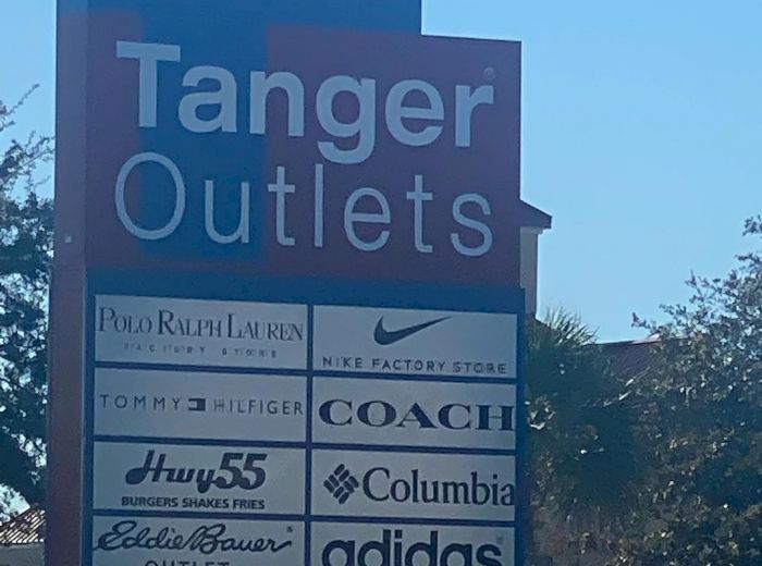 A sign for Tanger Outlets displaying brand names: Polo Ralph Lauren, Nike, Tommy Hilfiger, Coach, Hurley, Columbia, Eddie Bauer, and Adidas.