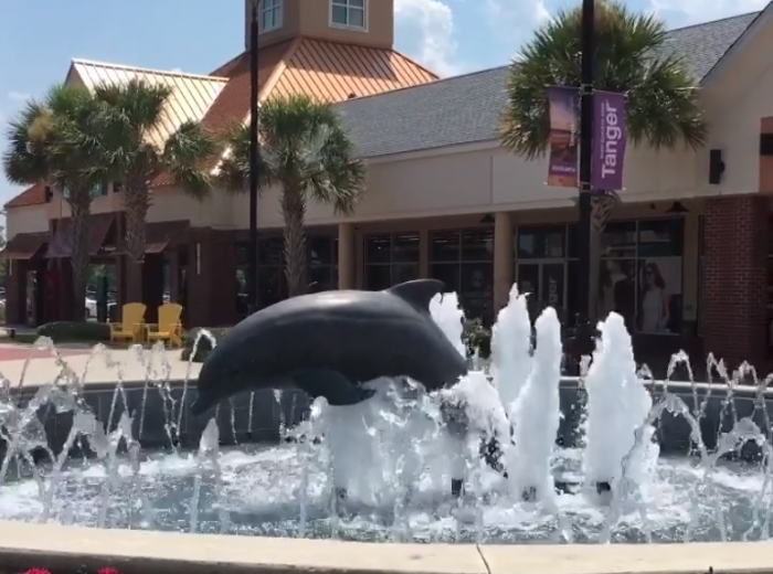 A dolphin statue in a water fountain is in front of a building. Palm trees and a purple banner are also present outside the building.