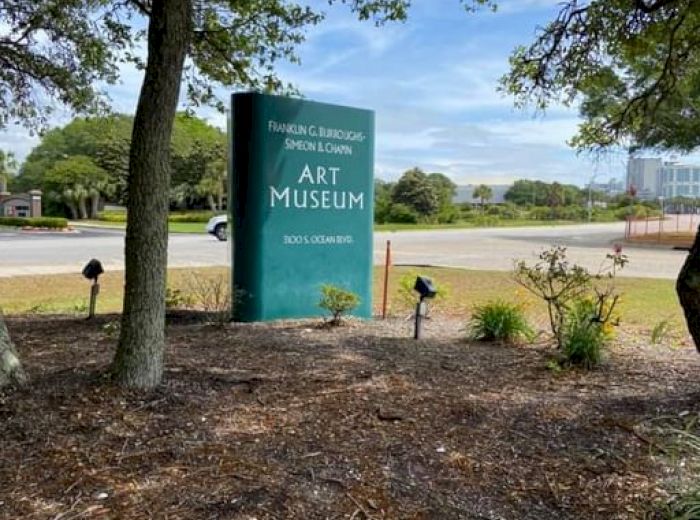 The image shows a green sign amidst trees and grass that reads "Art Museum." The background features a parking lot and buildings.