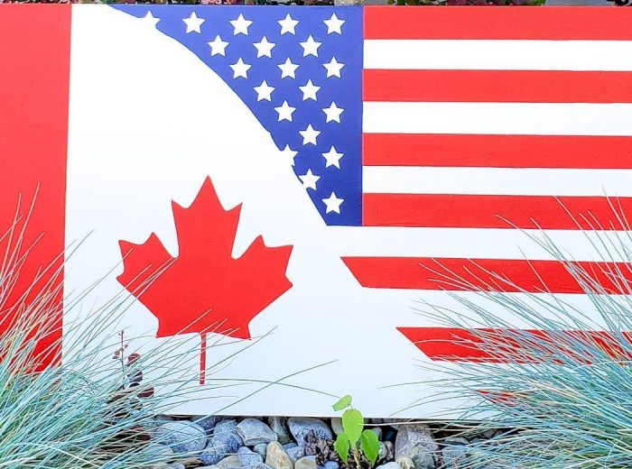 An artistic sign combines elements of the U.S. flag and Canadian flag, placed outdoors among plants and rocks, featuring a maple leaf and stars.