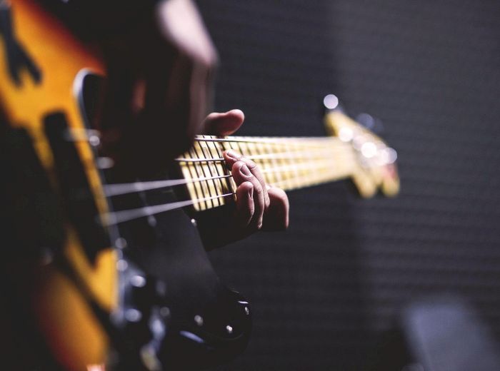 A close-up of a person playing an electric bass guitar, focusing on the fingers pressing the strings on the fretboard, with blurred background.