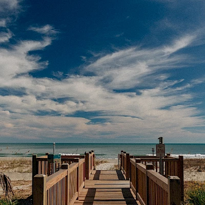 A wooden boardwalk leads to a sandy beach with clear blue skies and scattered clouds, overlooking the ocean.