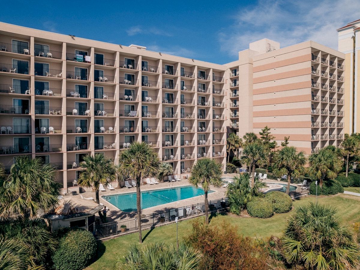 A multi-story hotel building with balconies, surrounded by palm trees, features an outdoor swimming pool and landscaped gardens.