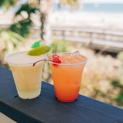 Two colorful cocktails, one yellow with a lime slice, and the other orange with ice and a cherry, placed on a railing with a beach view.