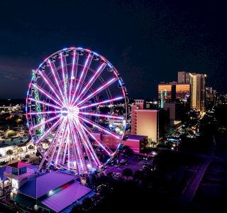 The image shows a Ferris wheel brightly lit with colorful lights at night, surrounded by city buildings and streets also illuminated.
