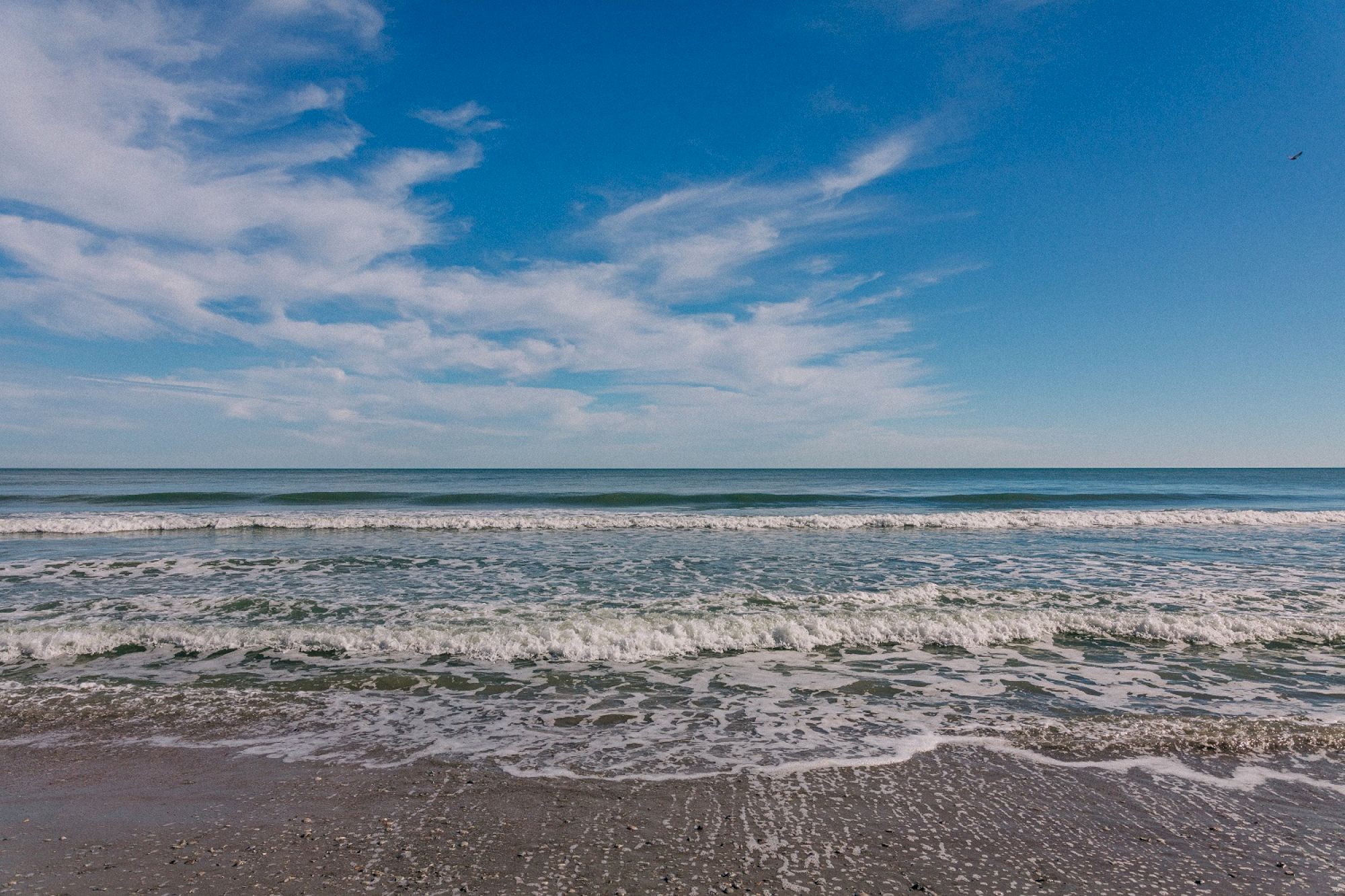 A serene beach scene with gentle waves rolling onto the shore under a partly cloudy blue sky, suggesting a peaceful coastal day.
