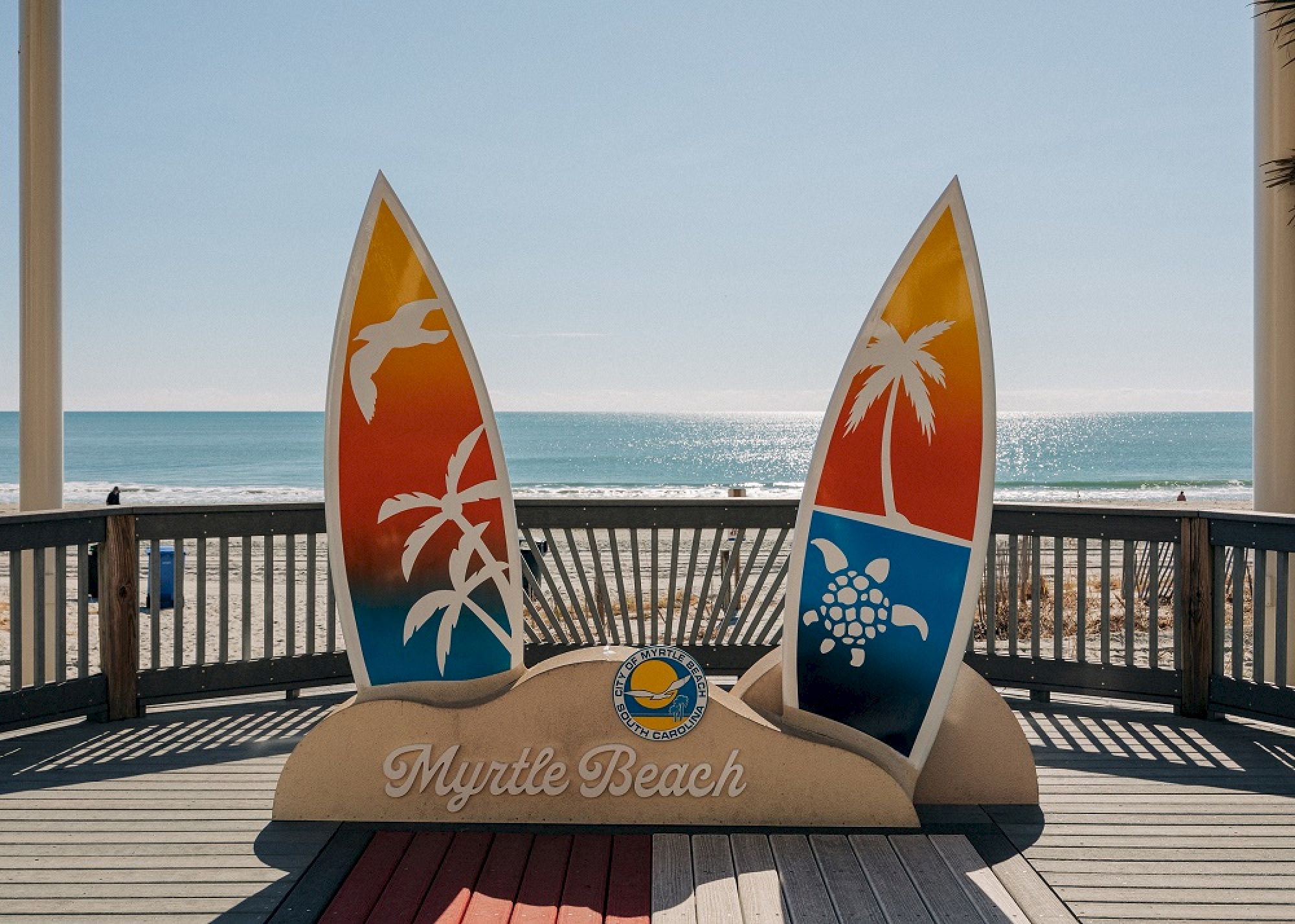 The image shows a Myrtle Beach sign in front of an ocean backdrop, featuring surfboard designs with tropical motifs on a wooden deck.