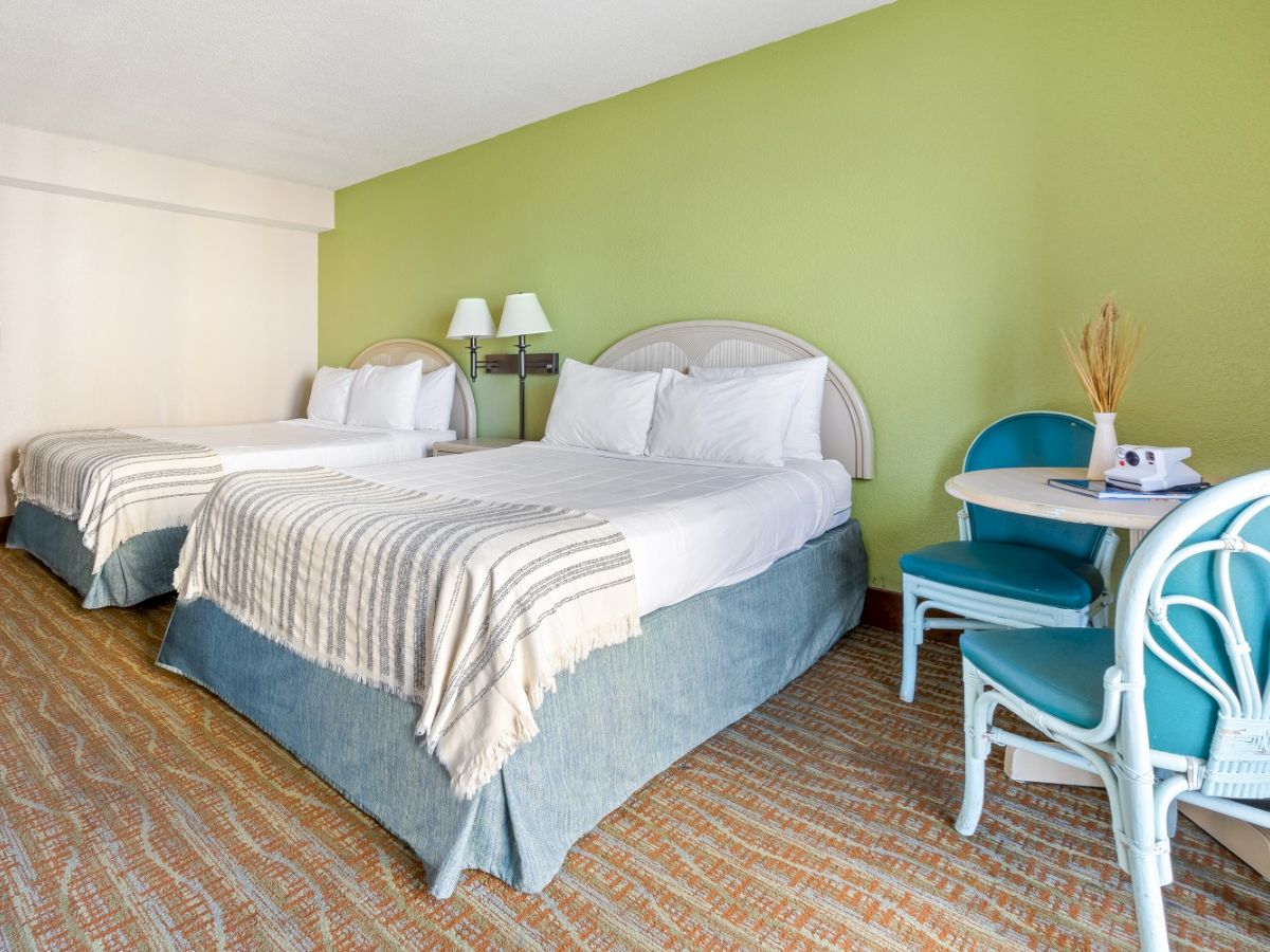 The image shows a hotel room with two double beds, a small table with two chairs, and light green walls, creating a cozy and inviting atmosphere.