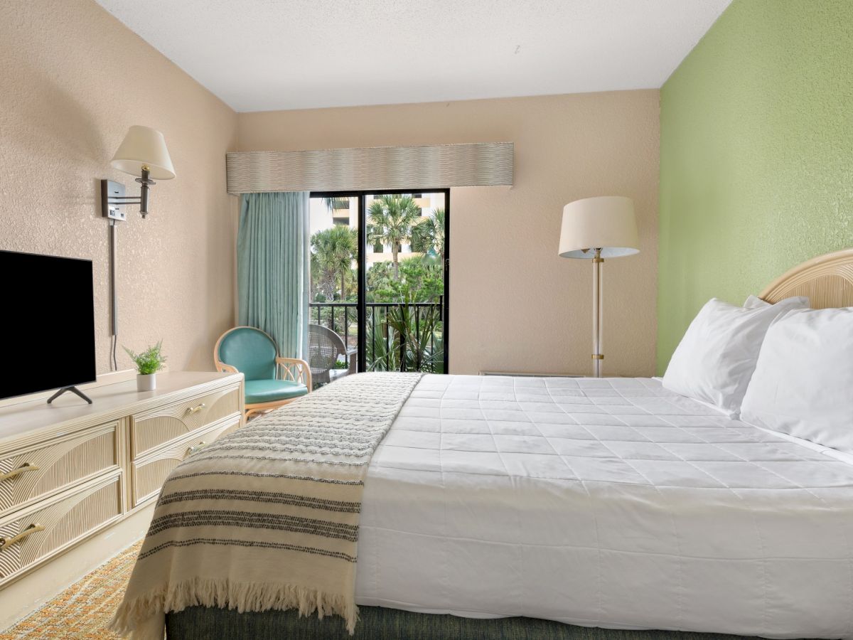 The image shows a bright hotel room with a large bed, TV, dresser, and a balcony offering a view of greenery outside.