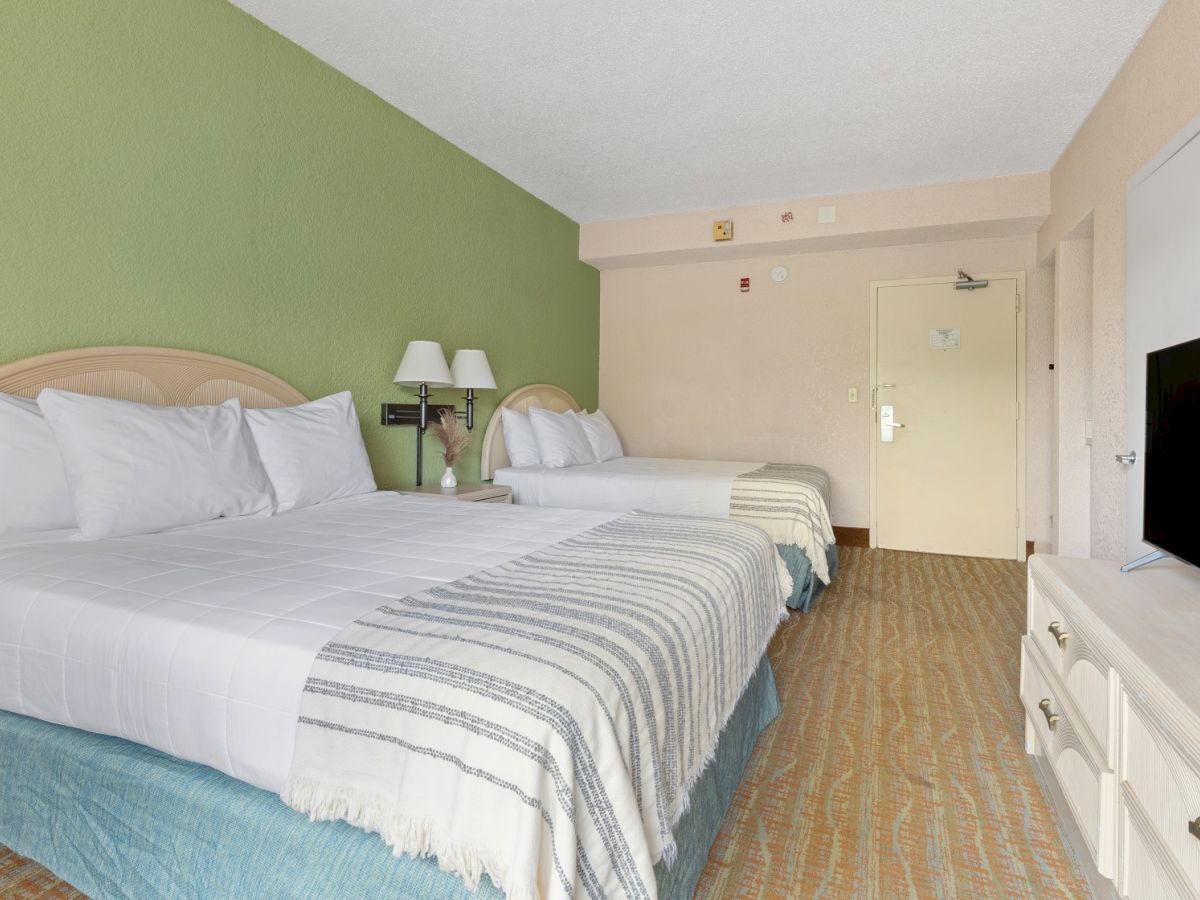 This image shows a hotel room with two beds, green accent wall, lamps, a TV on a dresser, and an open door at the far end of the room.
