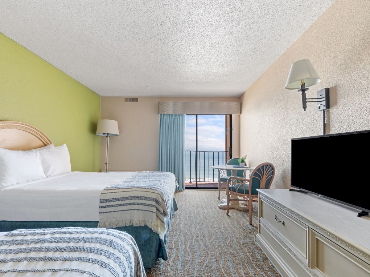 The image shows a hotel room with two beds, a flat-screen TV, a small table with chairs, and a sliding glass door opening to a balcony.