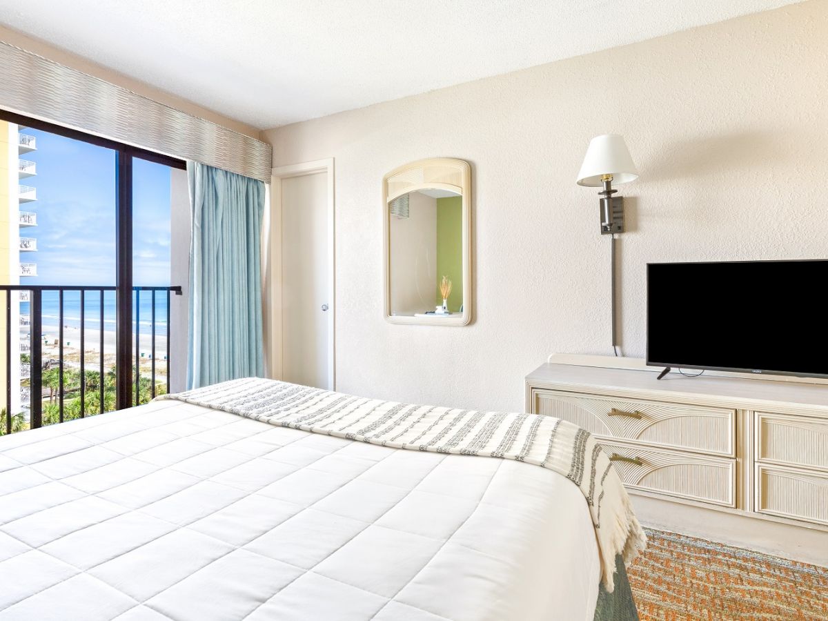 A bright hotel room with a bed, dresser, flat-screen TV, mirror, and a window that offers a view of the ocean and buildings outside.