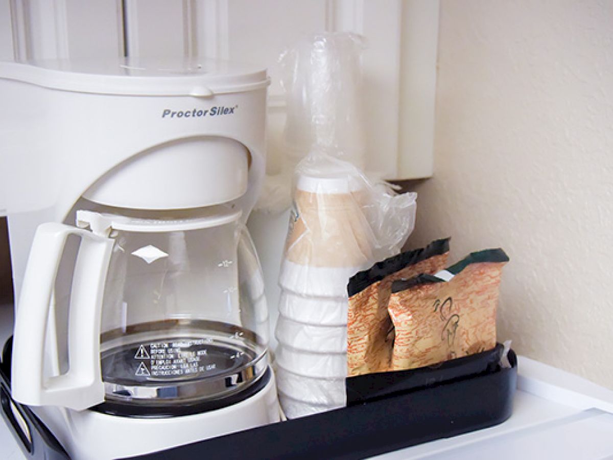 A coffee maker with a glass carafe sits beside disposable cups wrapped in plastic and packets of coffee grounds on a black tray in a kitchen nook.