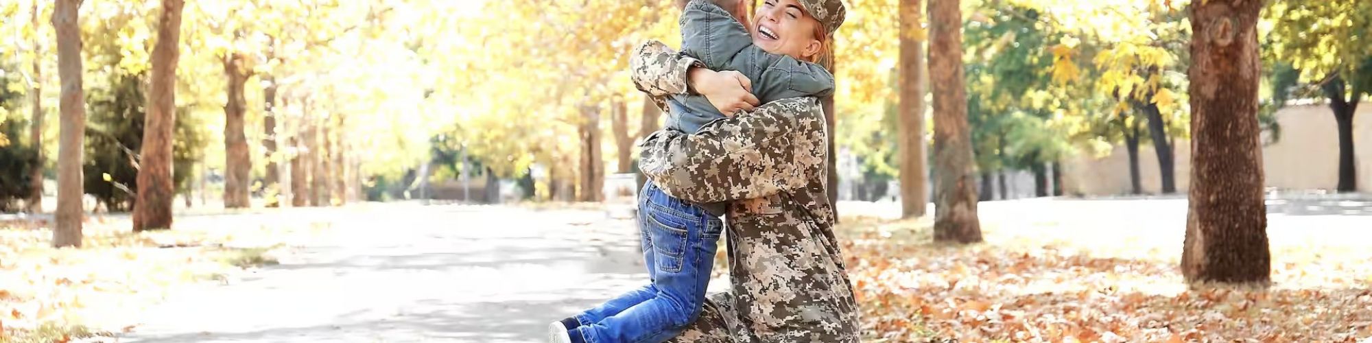 A person in military uniform hugs a child in an outdoor park, surrounded by trees and fallen autumn leaves.