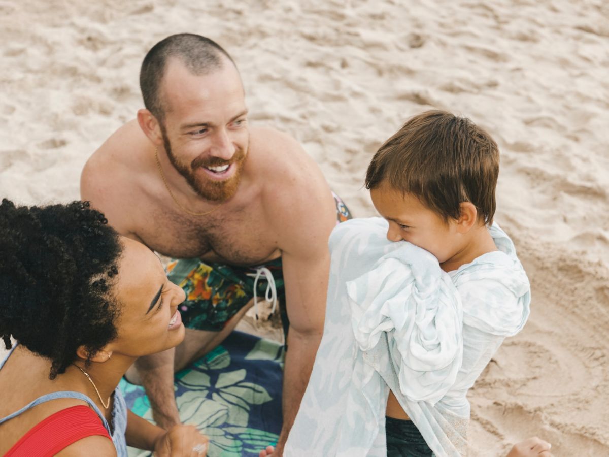 A man, woman, and child are enjoying their time together on a sandy beach, with the child playfully covering themselves with a towel.