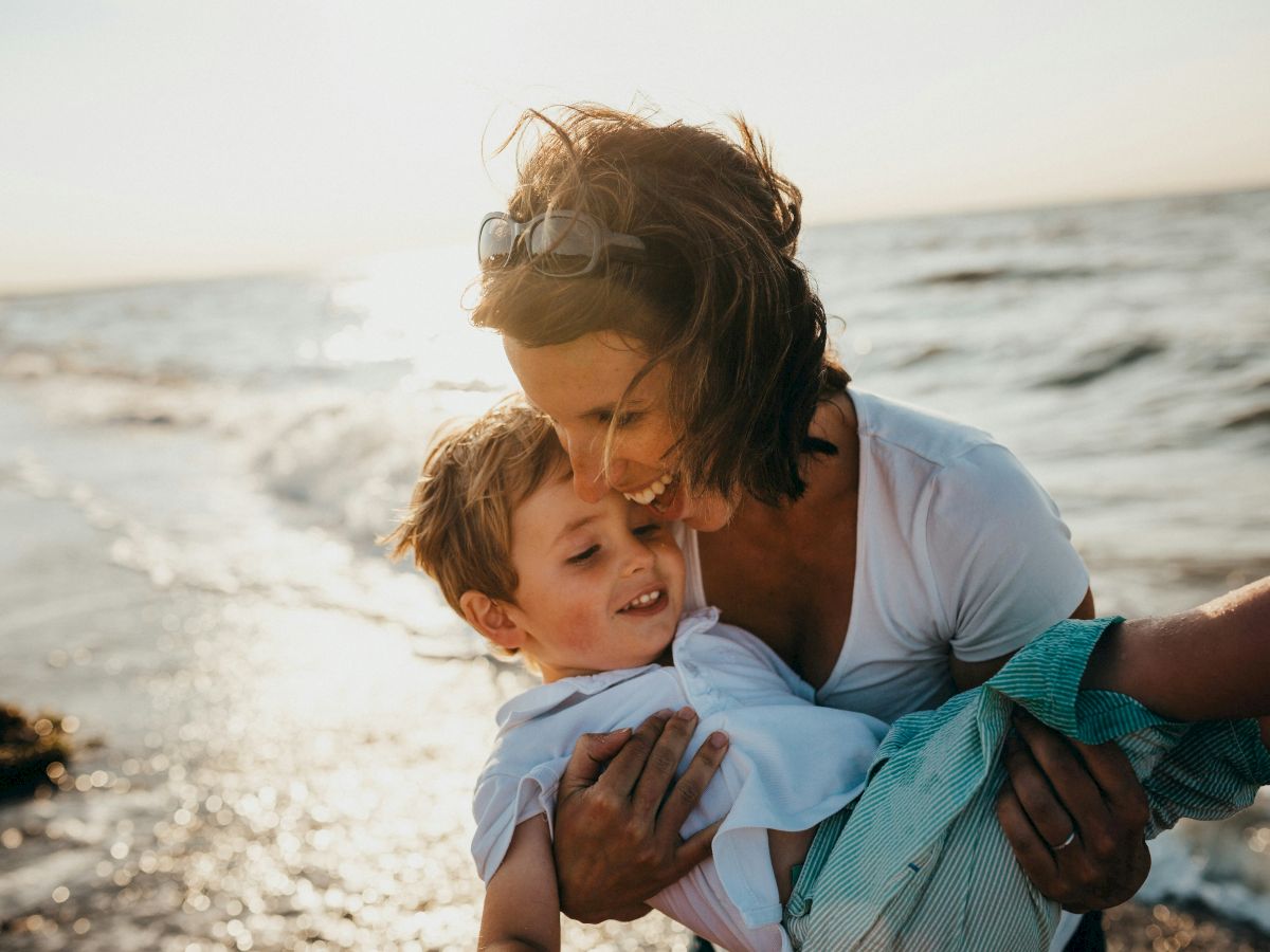 A woman is holding a child near the shore, both are smiling, with the ocean and a bright sky in the background.