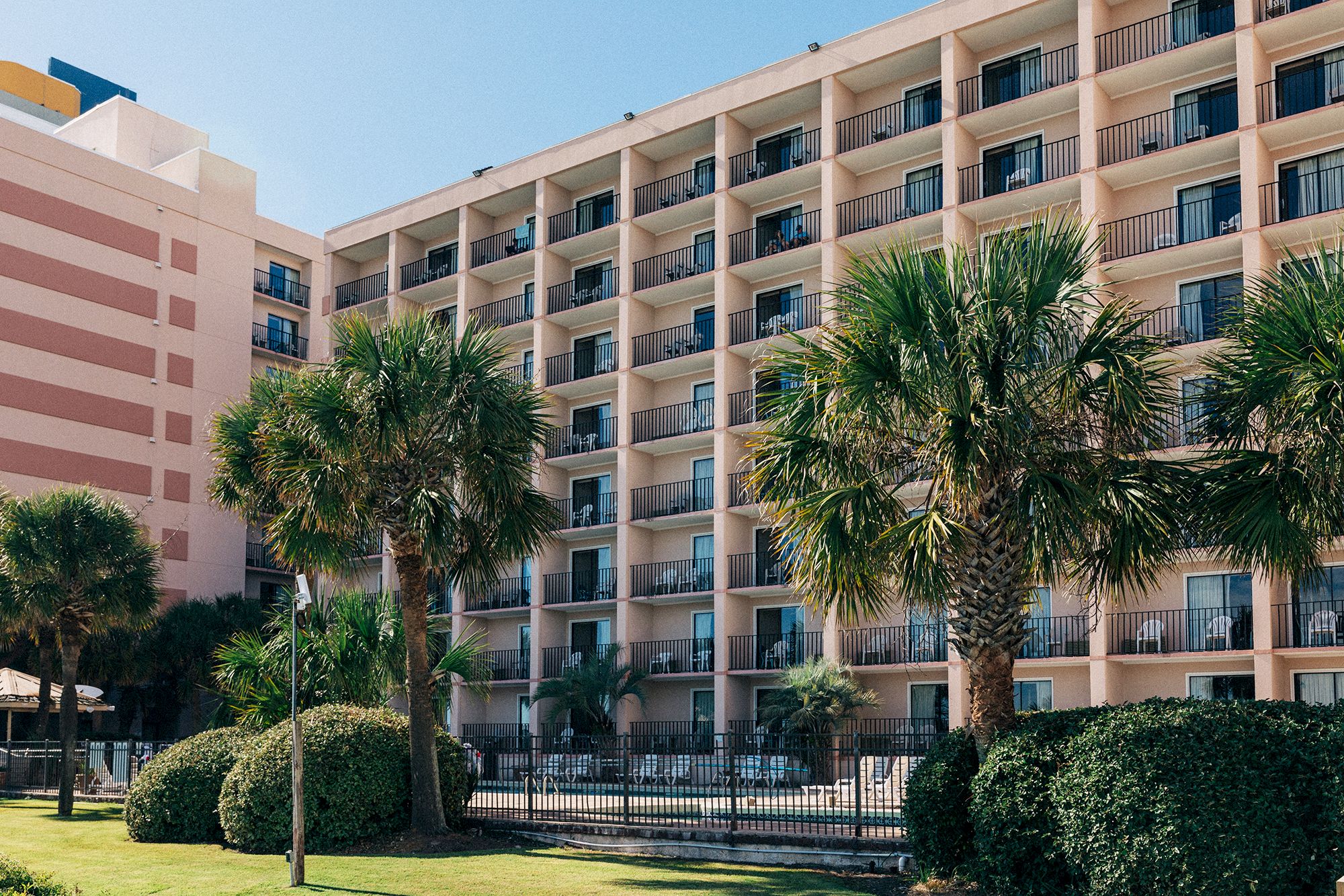The image shows a multi-story hotel with balconies, palm trees, and a fenced area, possibly for a pool, in the foreground.