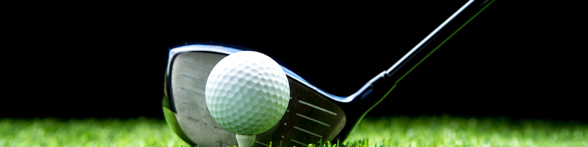 The image shows a golf club positioned next to a golf ball on a tee, set on green grass with a dark background.