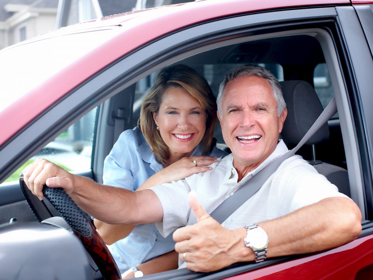 An older couple is sitting in a red car, smiling; the man is giving a thumbs up.