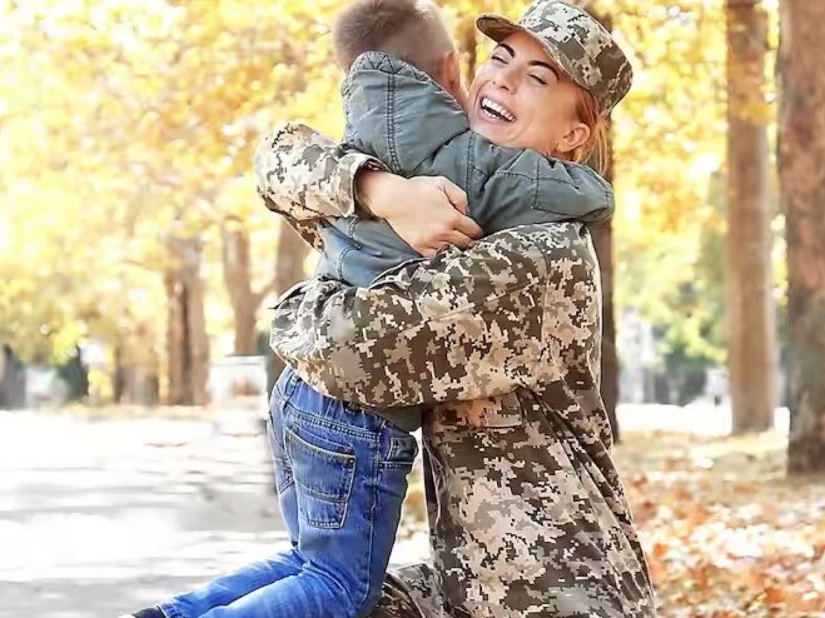 A person in camouflage uniform hugs a child outdoors in a park with autumn leaves in the background, both appear joyful and happy.